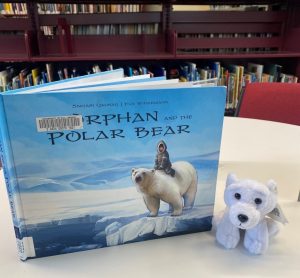 Finger puppet of Moka the Spirit Bear sits next to a picture book titled 'The Orphan and the Polar Bear'.